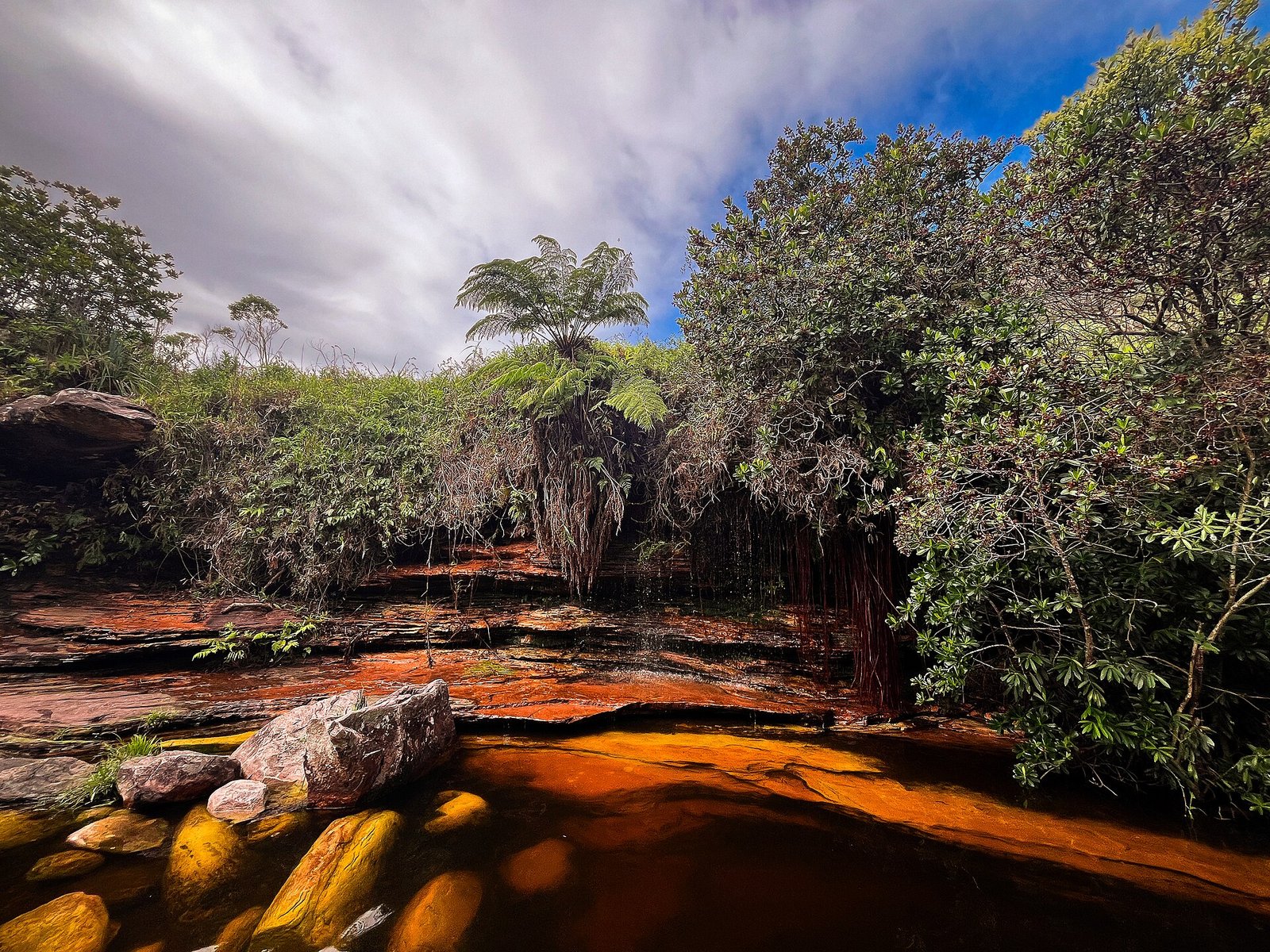 Poço de água avermelhada entre lajes de pedra e vegetação de mata, sob céu nublado