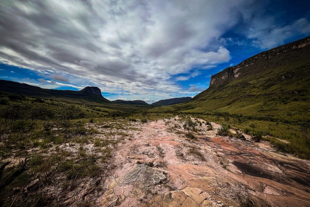 Trilha sobre lajedo de pedra em vale aberto, com morros e paredões ao fundo