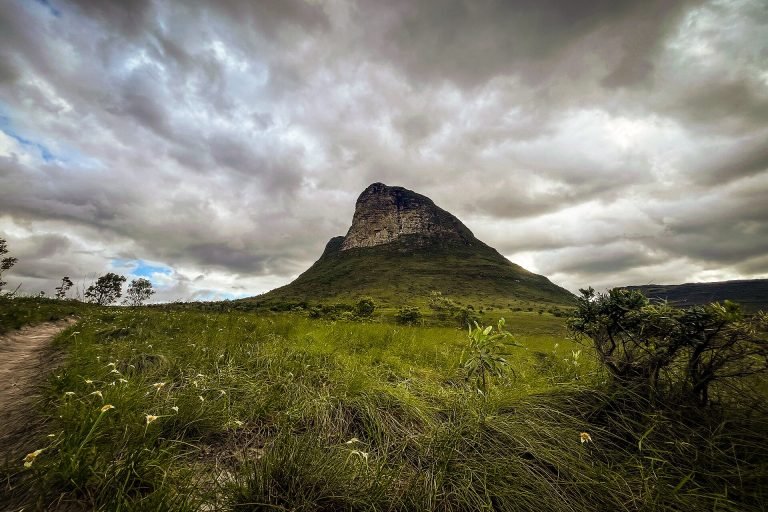 Morro de topo rochoso sobre campo de capim verde sob céu carregado