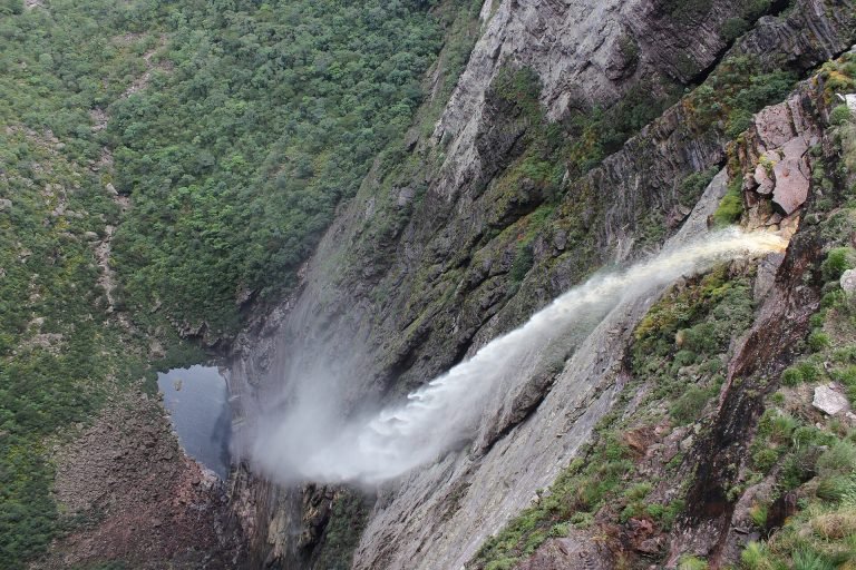 Queda d'água fina se desfazendo em névoa ao longo de um paredão de rocha, com o poço lá embaixo
