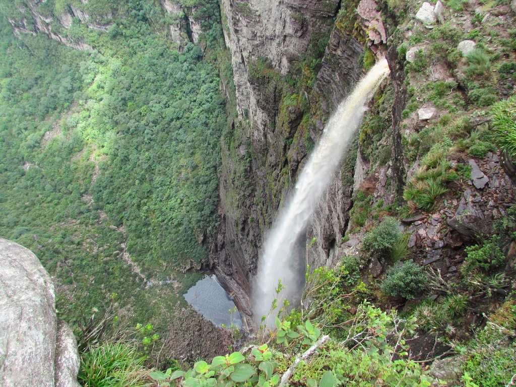 Cachoeira despencando do paredão rochoso coberto de vegetação, com poço escuro no fundo do cânion