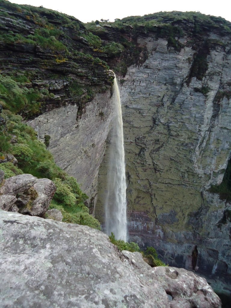 Fio de água caindo de um paredão cinza, visto da borda de pedra do mirante