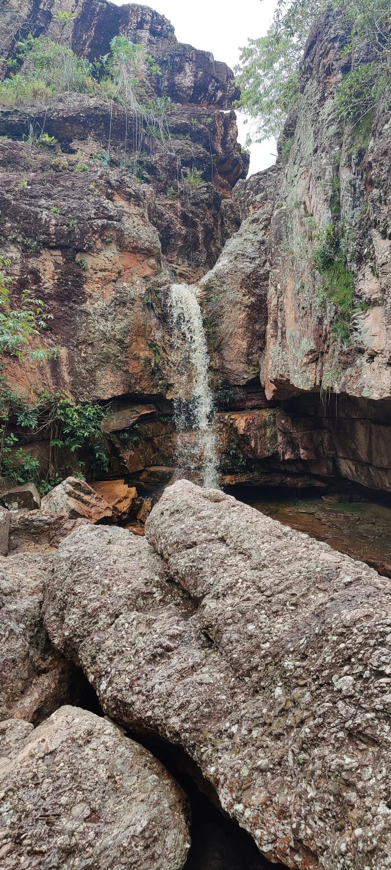Queda d'água fina entre paredões de rocha, com blocos de pedra em primeiro plano
