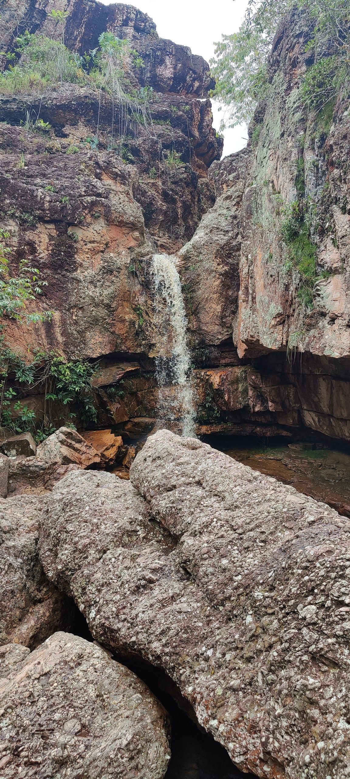 Queda d'água fina entre paredões de rocha, com blocos de pedra em primeiro plano
