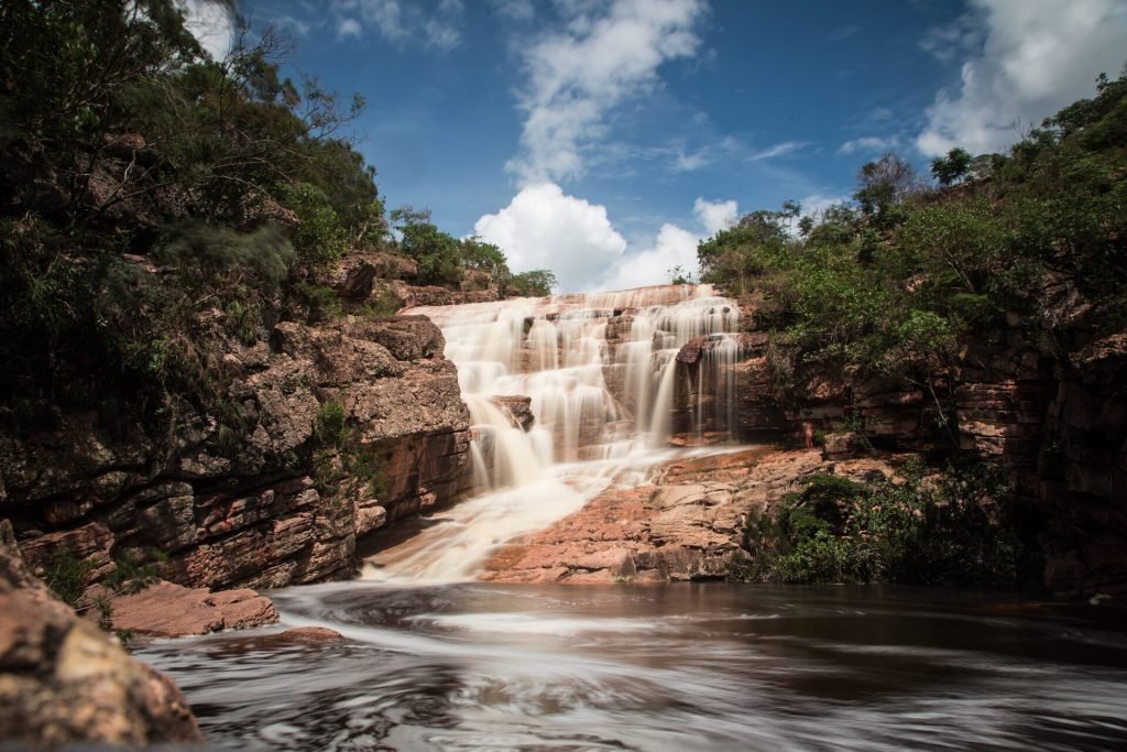 Água em longa exposição descendo degraus de pedra até o rio, entre mata