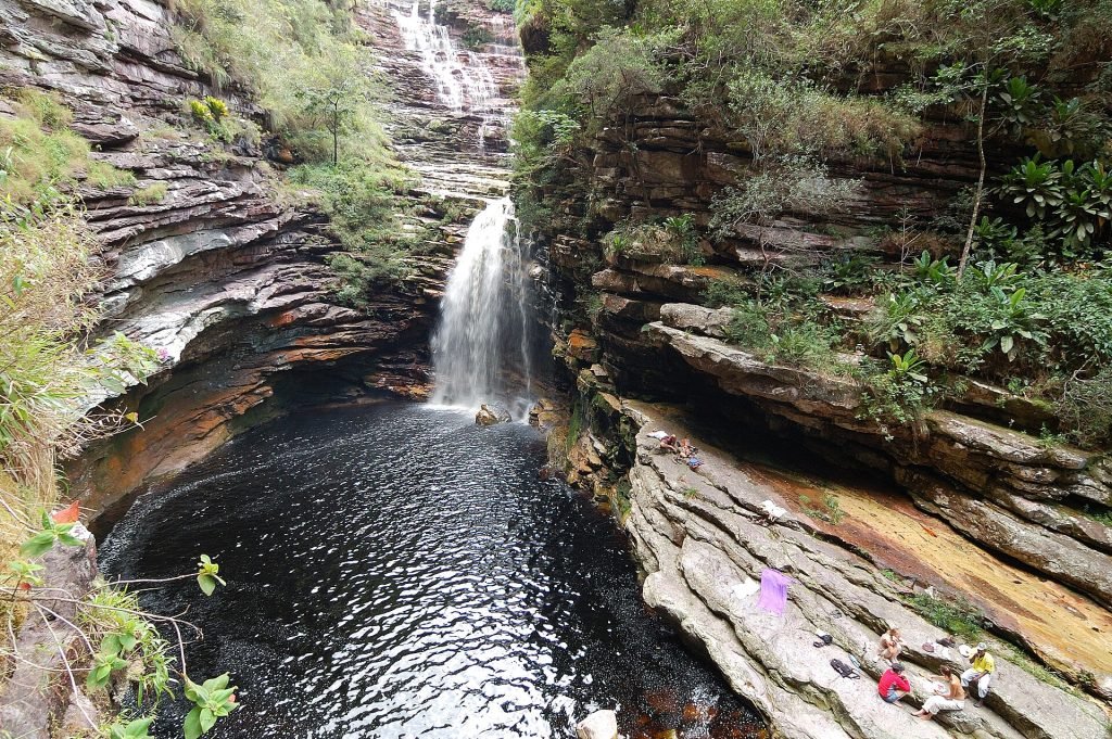 Cachoeira e poço escuro cercados por camadas de rocha e vegetação