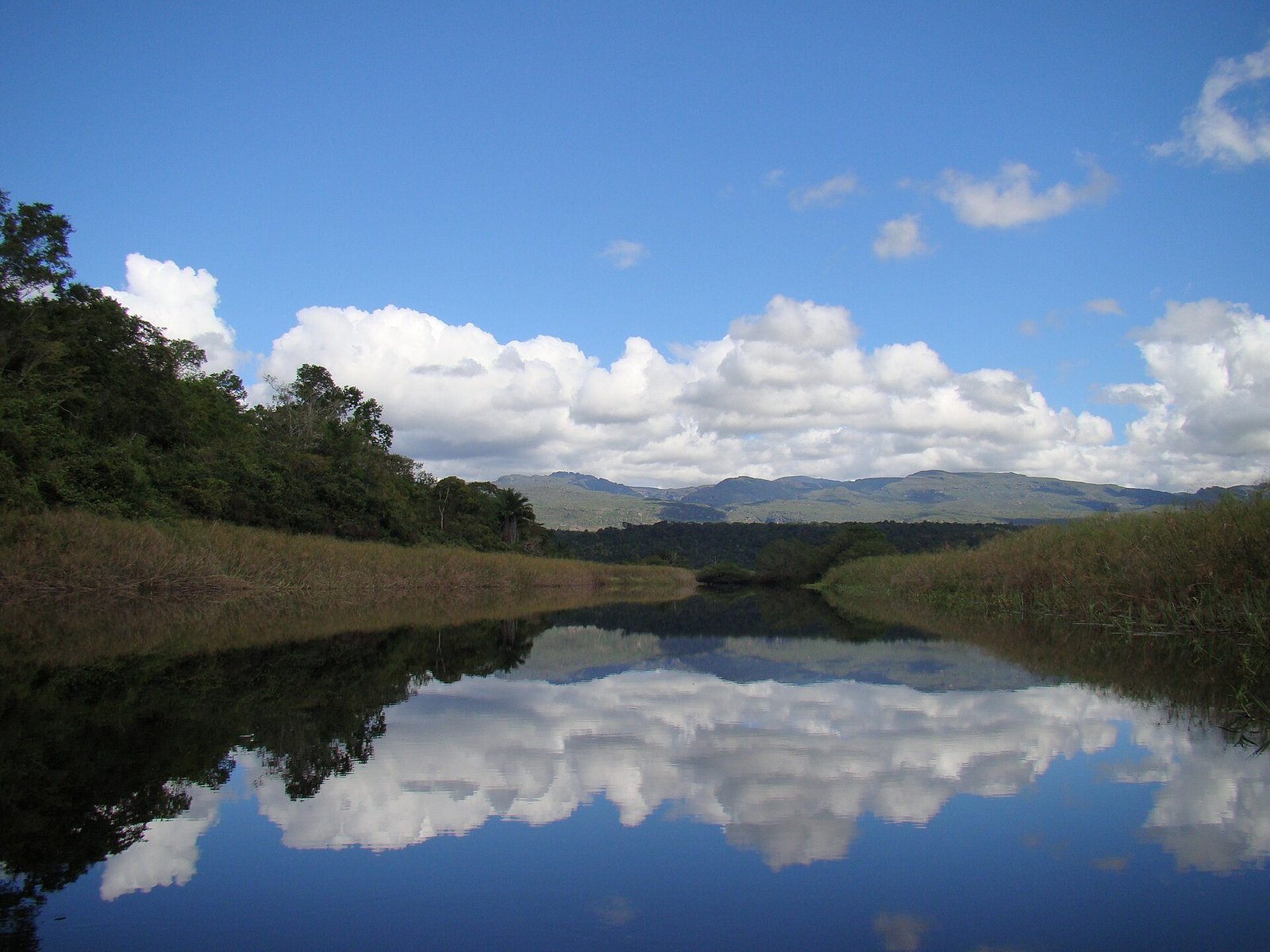 Rio de águas calmas refletindo céu e nuvens, entre vegetação alagada e serras ao fundo