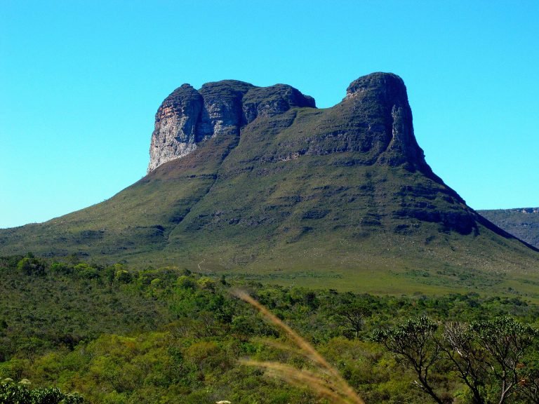 Morro com dois picos de rocha que lembram as corcovas de um camelo, sobre encosta verde e céu azul