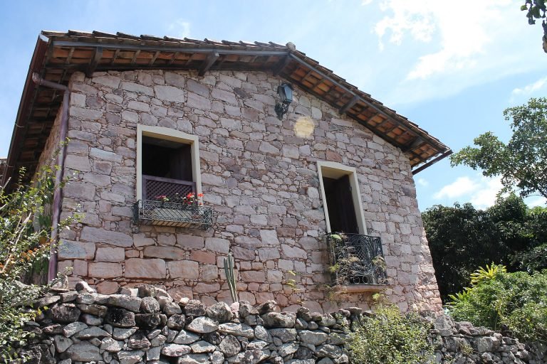 Casa de pedra com janelas de madeira e floreiras, cercada por muro de pedra e vegetação