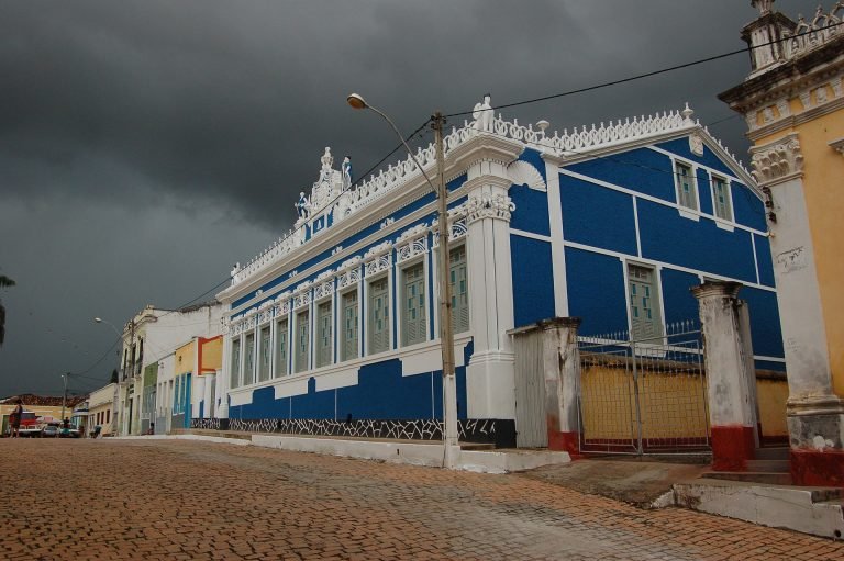 Casarão azul e branco de fachada ornamentada em rua de paralelepípedo, sob céu escuro de chuva