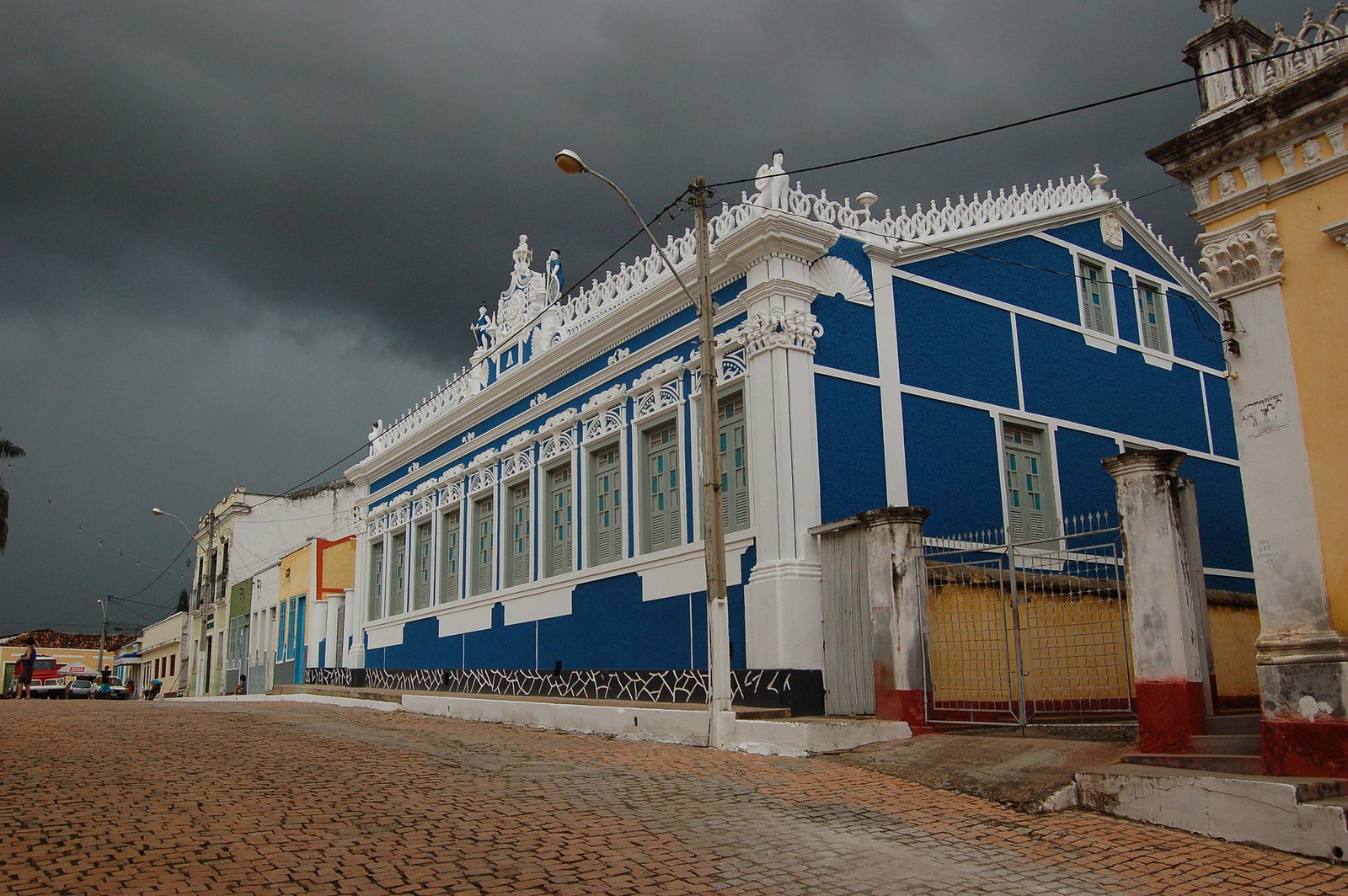 Casarão azul e branco de fachada ornamentada em rua de paralelepípedo, sob céu escuro de chuva