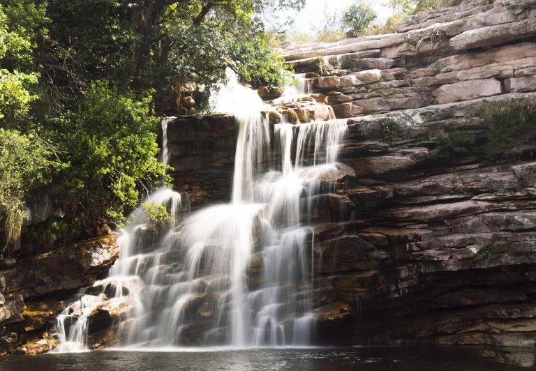 Cachoeira em degraus de rocha caindo em poço escuro, com mata no alto do paredão