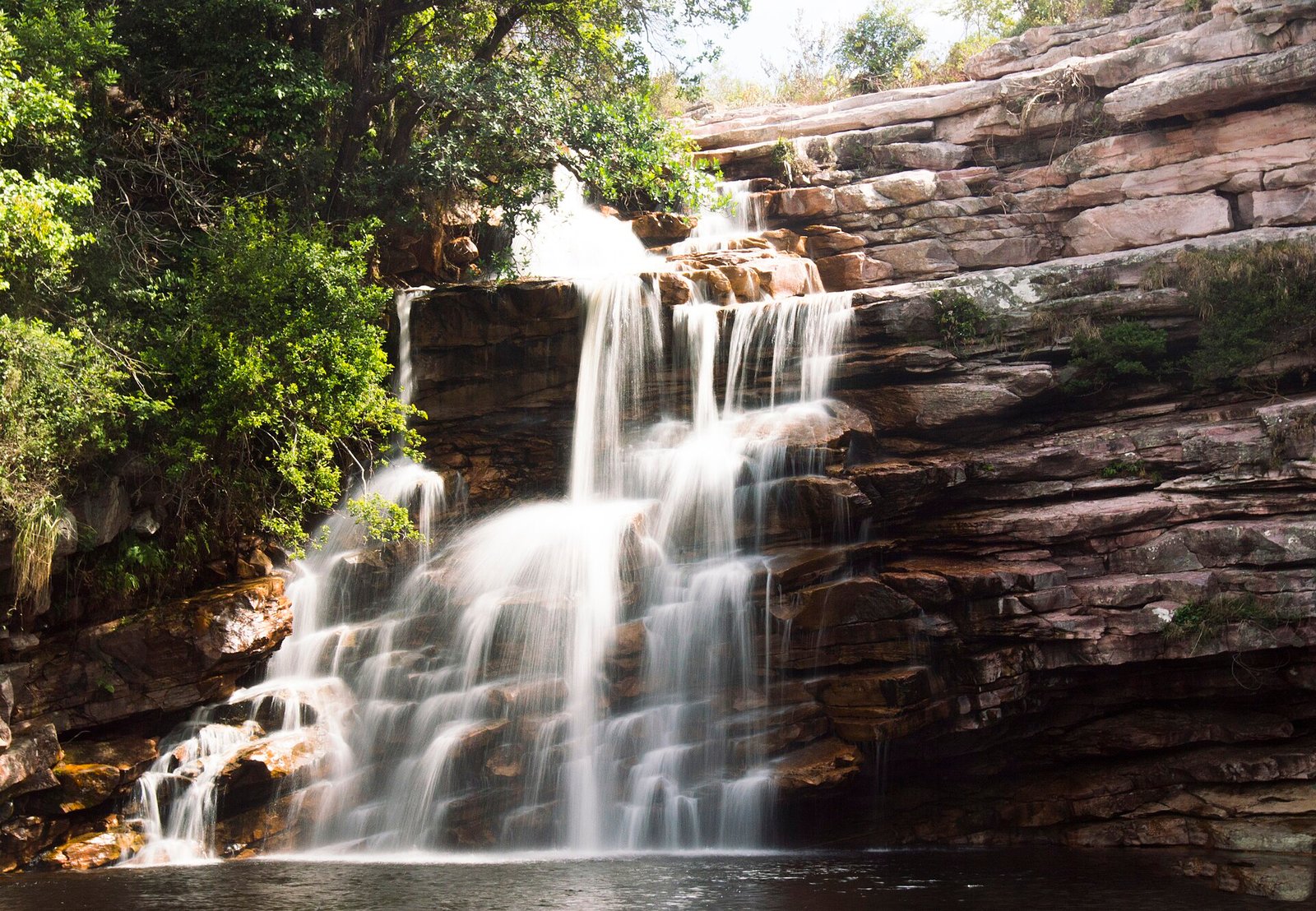 Cachoeira em degraus de rocha caindo em poço escuro, com mata no alto do paredão