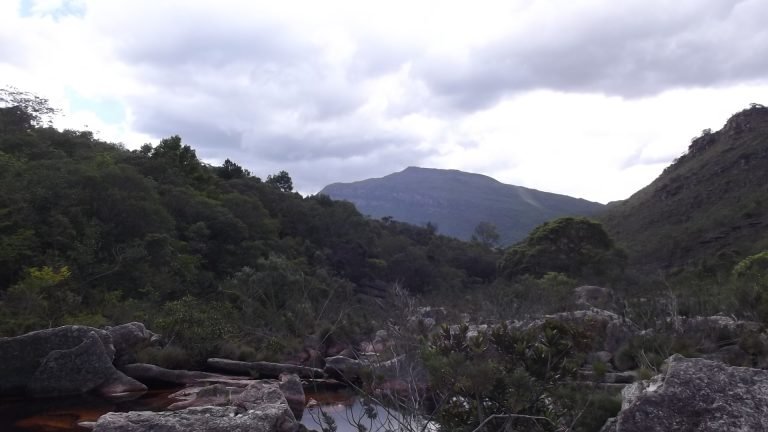 Poço de água escura entre rochas e vegetação de mata, com morro ao fundo sob céu nublado