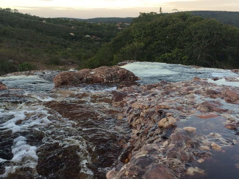 Água escura do rio correndo sobre rochas avermelhadas, com morros ao fundo