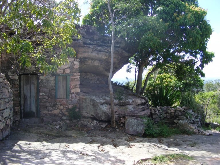 Casa de pedra com portas de madeira encaixada sob um grande bloco de rocha