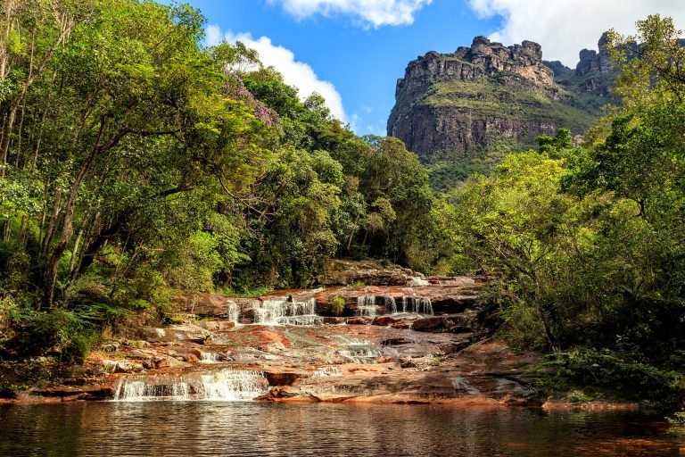 Pequenas quedas d'água em rio de pedras avermelhadas, com o Morro do Castelo ao fundo